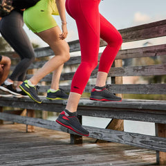 Women climbing wooden steps outdoors wearing GORUCK athletic shoes in wolf grey with red accents and colorful workout leggings