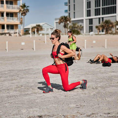 Woman in red athletic wear lunging on beach while wearing black GORUCK backpack during outdoor workout