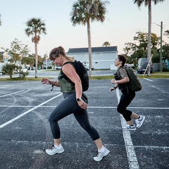 Two women in athletic wear doing lunges outdoors wearing GORUCK backpacks in a parking lot with palm trees