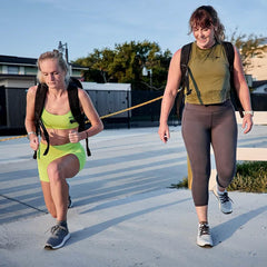 Two women exercising outdoors wearing GORUCK backpacks, one in green activewear lunging and the other in gray leggings stepping