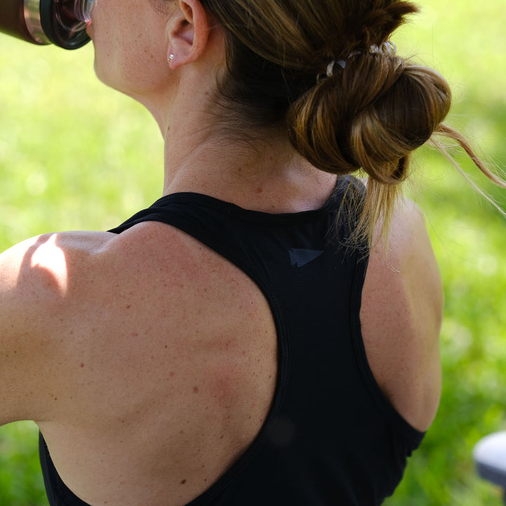 A woman with her hair in a bun drinks from a bottle outdoors, wearing the Women’s Racerback Tank - ToughMesh in sleek black premium fabric.