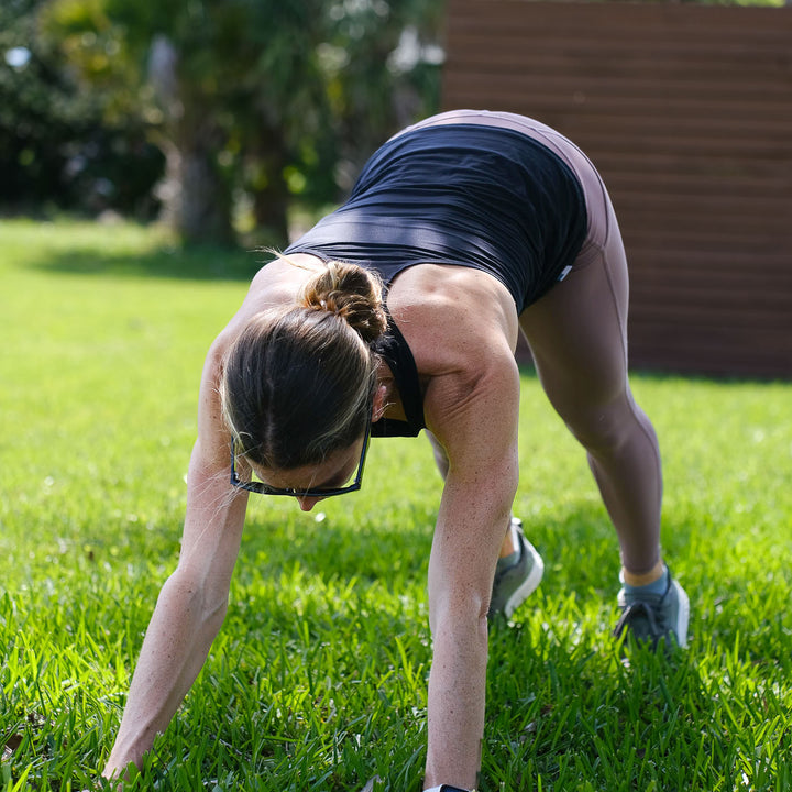 A woman in a crawling pose exercises outdoors on grass, wearing glasses and the Women’s Racerback Tank - ToughMesh with athletic clothes.