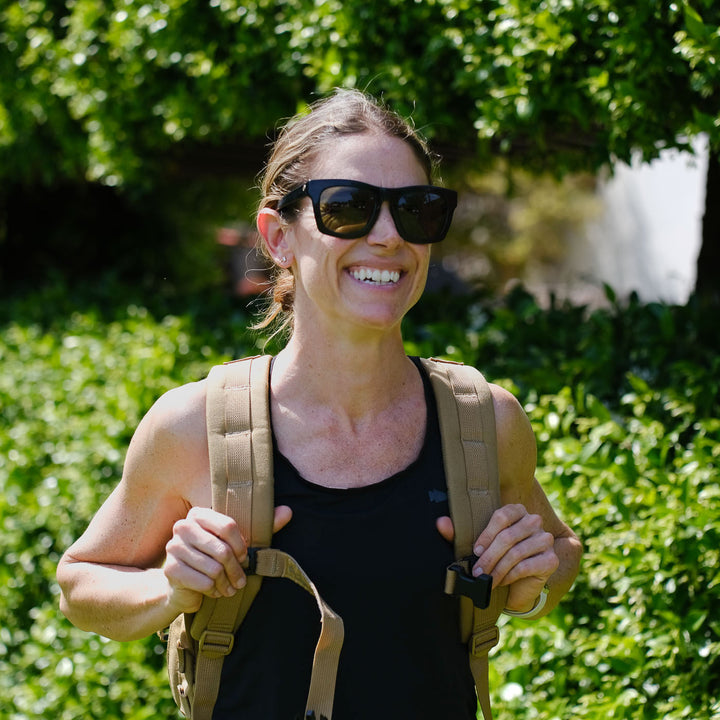 A woman wearing sunglasses and a backpack smiles outdoors with green foliage behind her, showing off her Women’s Racerback Tank - ToughMesh made from premium fabric.