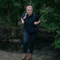 Woman with pink braided hair hiking in black fleece vest and backpack in forest trail