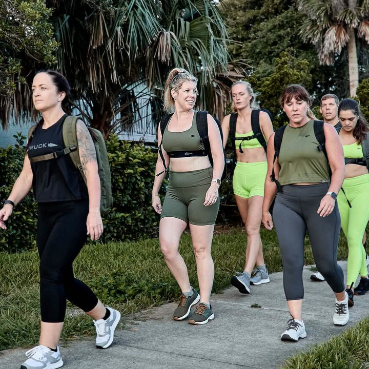 A group of women dressed in athletic gear strolls along a sidewalk surrounded by greenery. Some sport matching green outfits made from GORUCK's ToughFlex fabric, perfect for their fitness activity. Others are wearing GORUCK’s Women's Biker Shorts - ToughFlex, ensuring comfort and durability on their energetic walk.