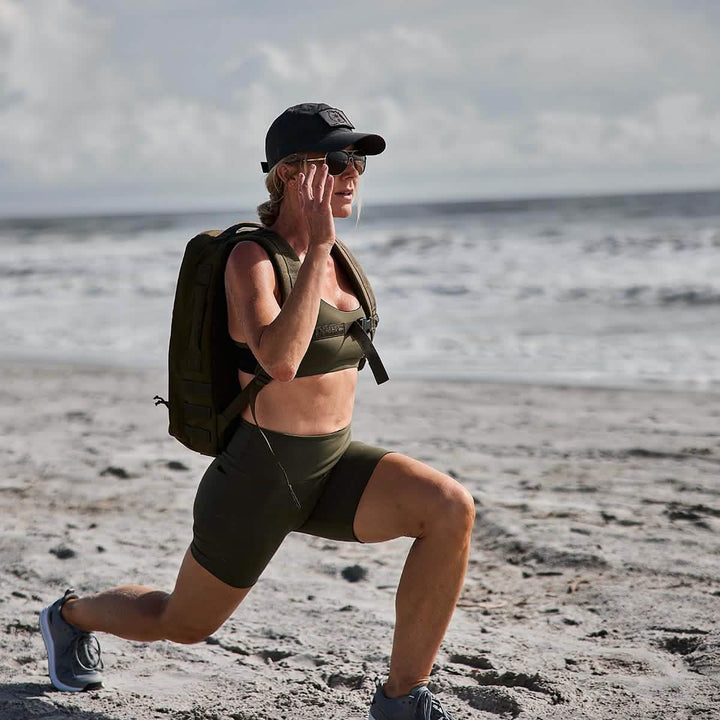 A person in athletic gear, sporting a cap and sunglasses, performs lunges on a sandy beach with ocean waves in the background. Clad in a sports bra and GORUCK's Women’s Biker Shorts made from ToughFlex fabric, they carry a backpack, prepared for any adventure.