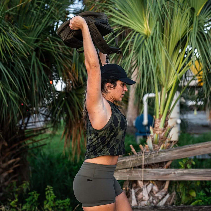 A person wearing a black cap and a camouflage tank top is exercising outdoors, lifting a bag overhead. They are clad in GORUCK's Women’s Squat Shorts made from ToughFlex Fabric, standing near tall palm trees with a wooden fence in the background.