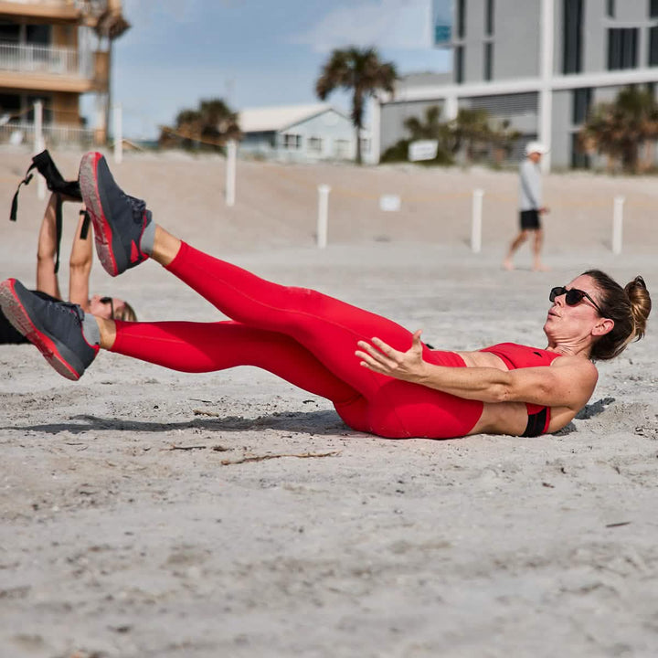 A woman wearing high-waisted GORUCK Women’s Training Leggings Pocket - ToughFlex is seen doing a core exercise on the beach, lifting her legs and upper body off the sand. Made from ToughFlex fabric, her leggings are ideal for active pursuits. Nearby, other people are exercising, with buildings and palm trees visible in the background.