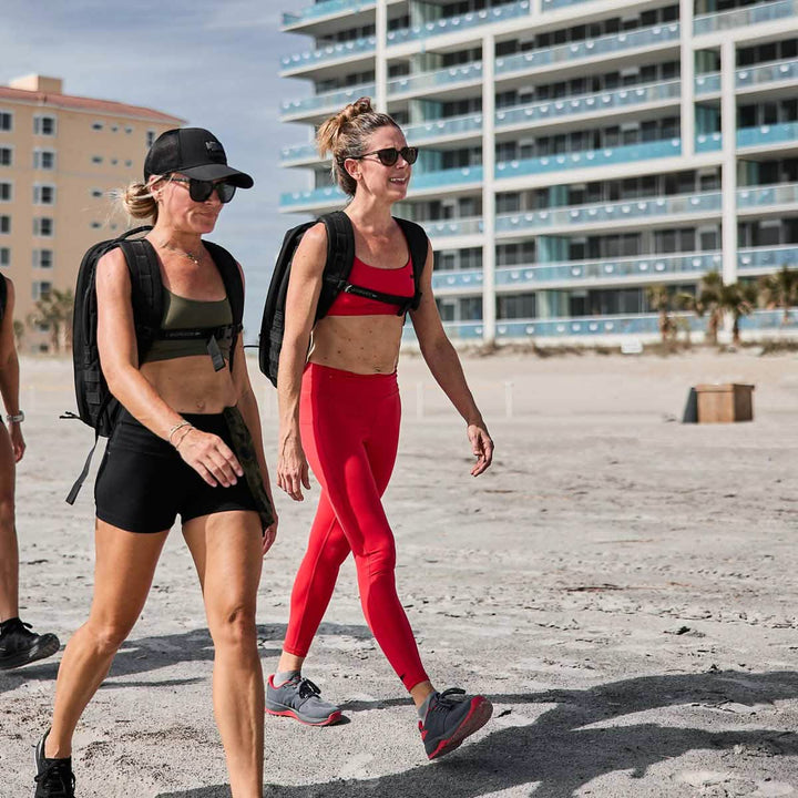 Two women in athletic gear, sunglasses, and backpack weights stroll along a sandy beach. One sports high-waisted leggings and a cap, while the other wears red GORUCK Women’s Training Leggings Pocket crafted from ToughFlex fabric. A multi-story building looms in the background.