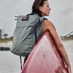 Woman carrying a gray GORUCK backpack with American flag patch, holding a red surfboard on a beach