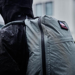 Close-up of waterproof GORUCK rucksack with water droplets on fabric, worn over black jacket in rainy setting