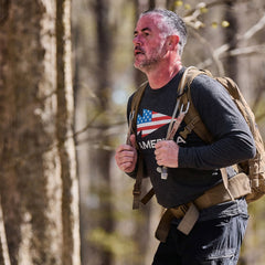 A man hikes in the woods wearing the Men's America Long Sleeve - Tri-BlendX and a backpack with an American flag design, looking focused.