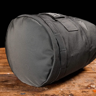 A Brick Bag from GORUCK, designed in a gray cylindrical shape akin to military gear, rests on a rustic wooden surface. Its durable woven texture and convenient top handle are highlighted against a solid black background.