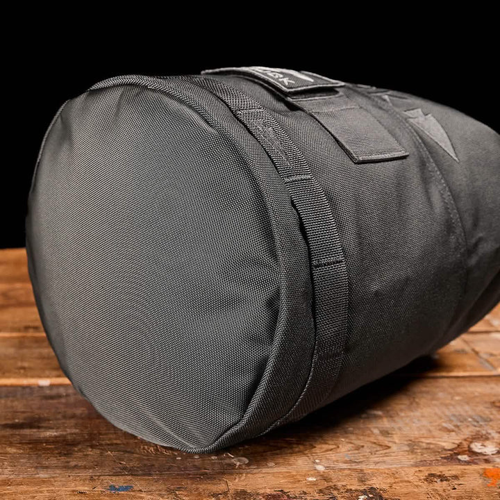 A Brick Bag from GORUCK, designed in a gray cylindrical shape akin to military gear, rests on a rustic wooden surface. Its durable woven texture and convenient top handle are highlighted against a solid black background.