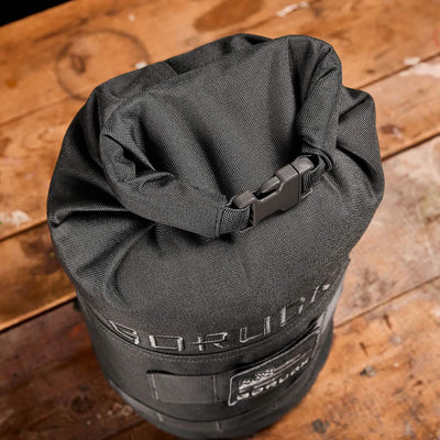 A close-up of a GORUCK Brick Bag (5-50 LBS) in green, showcasing its rolled top secured by a black buckle. It rests on a rustic wooden surface, embodying versatile training essentials, with the brand partially visible on the side of this tactical gear piece.