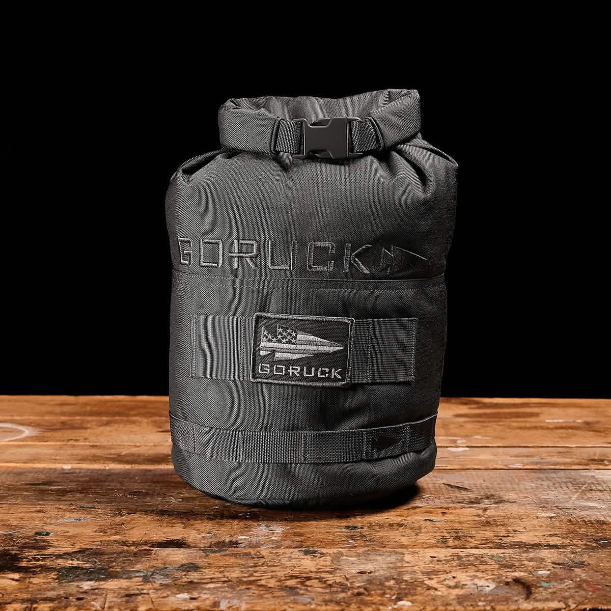 A black Brick Bag by GORUCK, inspired by military gear, rests on a wooden surface against a black background. It features a flag patch with "GORUCK" embroidered above it. This versatile training companion has a roll-top closure secured with a buckle.