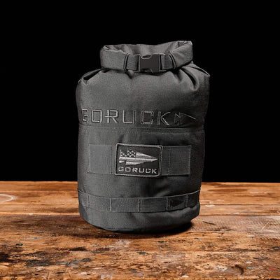 A black Brick Bag by GORUCK, inspired by military gear, rests on a wooden surface against a black background. It features a flag patch with "GORUCK" embroidered above it. This versatile training companion has a roll-top closure secured with a buckle.