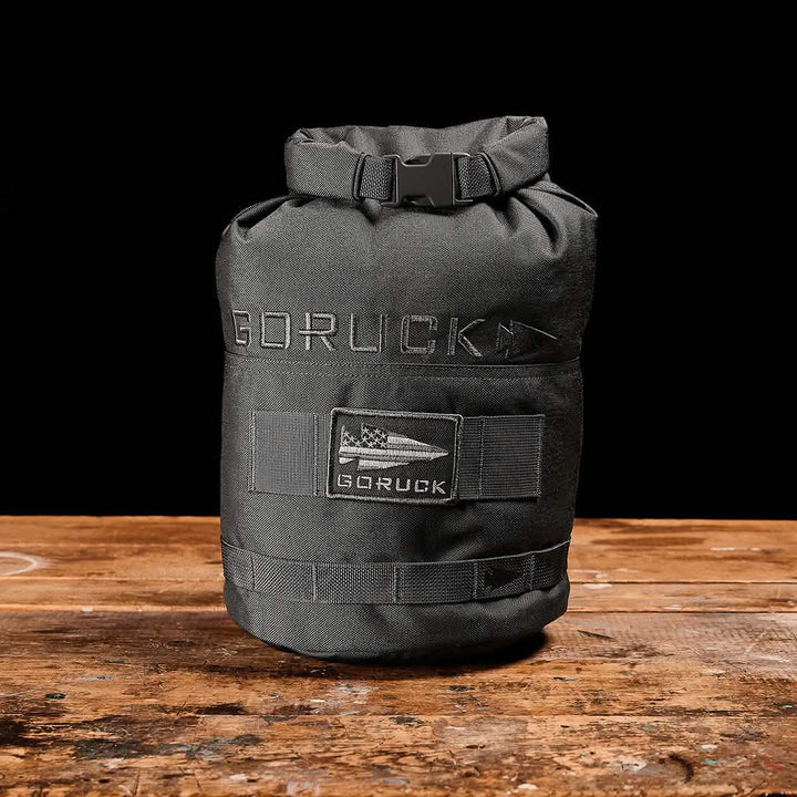 A black Brick Bag by GORUCK, inspired by military gear, rests on a wooden surface against a black background. It features a flag patch with "GORUCK" embroidered above it. This versatile training companion has a roll-top closure secured with a buckle.