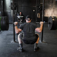 Man wearing black cap and athletic clothes performing heavy barbell squat in gym with black walls and workout equipment