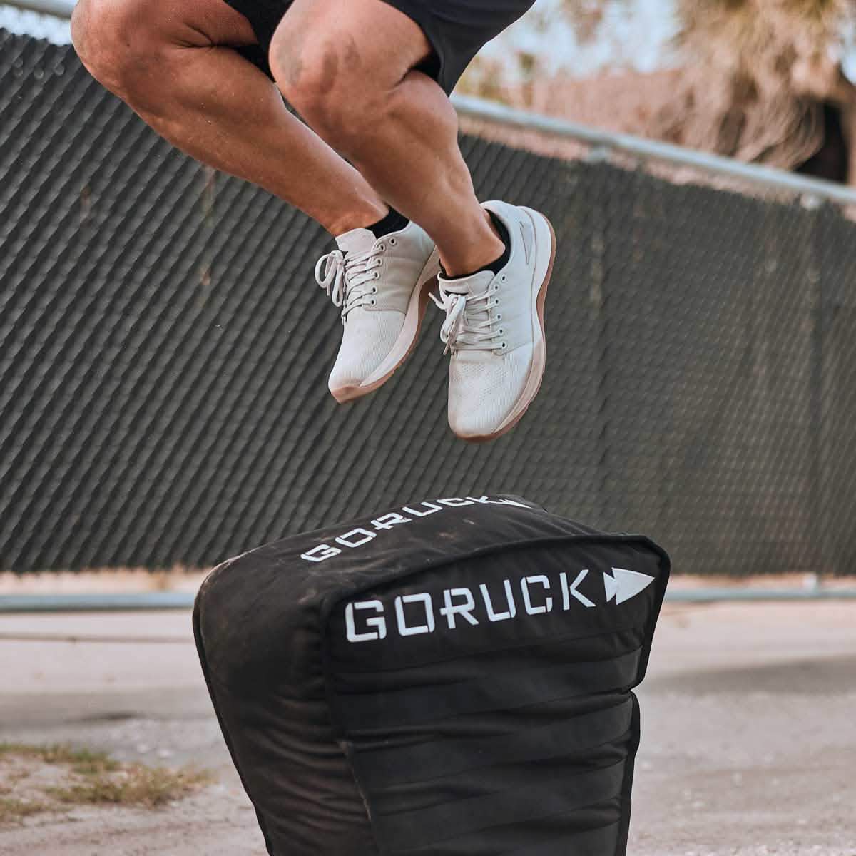A person wearing GORUCK's Men's Ballistic Trainers in Lunar Rock + Gum with a Silver Reflective Spearhead is jumping over a black GORUCK sandbag outdoors, set against a chain-link fence and the sky. The image captures the lower half of their athletic leap, showcasing the strength of CORDURA Ballistic Nylon and 3X Support in their footwear.