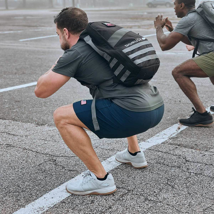 Two men engage in functional fitness exercises outside on a foggy day, squatting with backpacks that highlight their 3X Support and CORDURA Ballistic Nylon gear. The man in the foreground sports a gray shirt and blue shorts, while the man behind wears green. Both are wearing GORUCK Men's Ballistic Trainers in Lunar Rock + Gum with a Silver Reflective Spearhead as they work out on a marked parking lot.