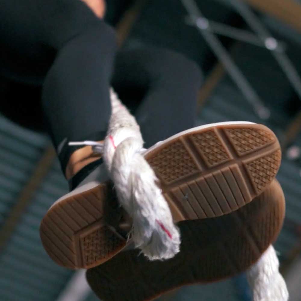 Close-up of a person’s shoes gripping a thick rope while climbing indoors.