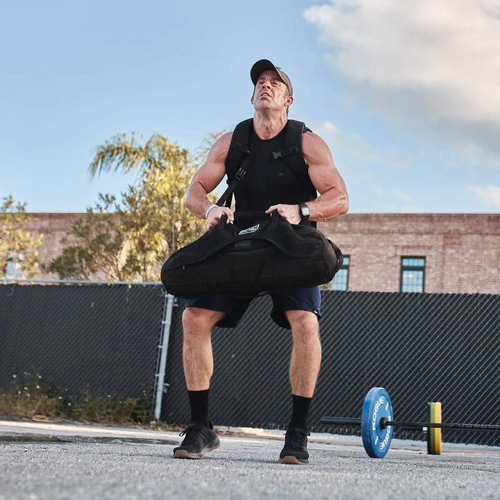 A muscular man sporting GORUCK's Men's Ballistic Trainers in Black + Gum with a Black Reflective Spearhead is lifting a heavy sandbag outdoors. He wears a cap and tactical vest, while a weight plate and chain-link fence are visible in the background, along with trees and a building under the clear sky.