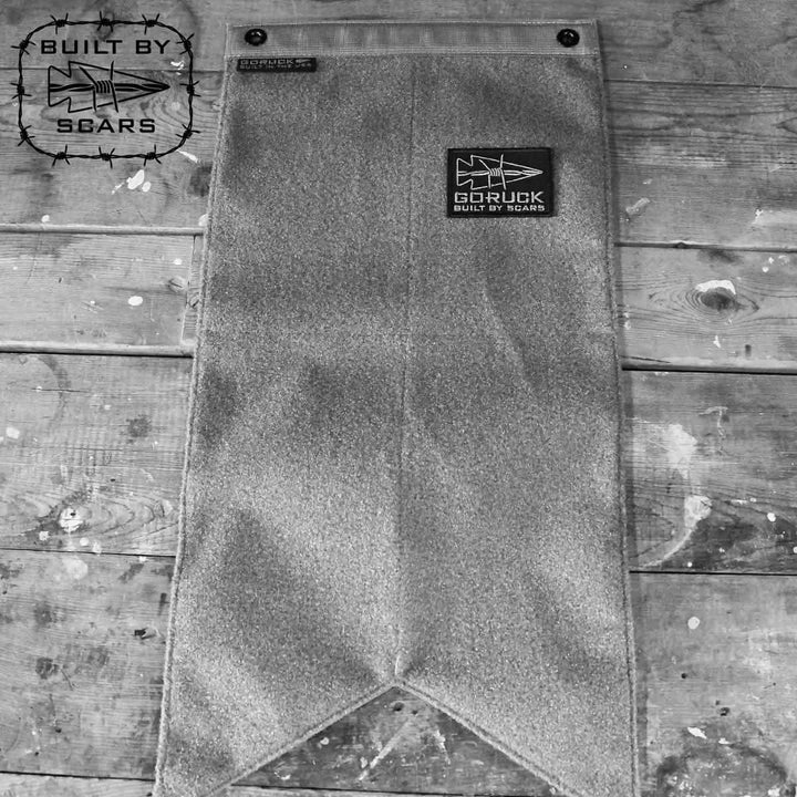 Black and white image of a GORUCK Banner Patch Board on a worn wooden surface. The velcro patch panel showcases a star and the text BUILT BY SCARS, alongside another small patch with a similar design, creating a rustic ambiance.