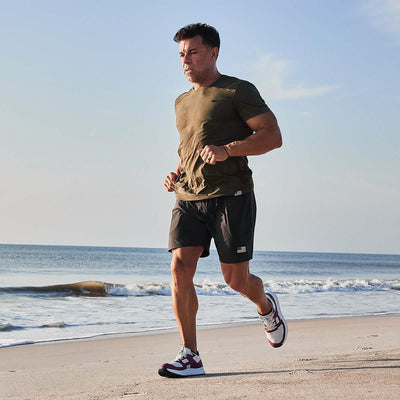 A man jogs on a sandy beach by the ocean under a clear blue sky, capturing the essence of a Men's Rough Runner - Baton Rouge. He wears a green T-shirt, black shorts, and sneakers from GORUCK. The waves gently lap at the shore in the background.