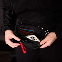 A person in a black jacket and pants holds a GORUCK Belt Bag with adjustable webbing. The sleek fanny pack is unzipped, showing a smartphone, wallet, and red multitool inside, keeping their gear organized.