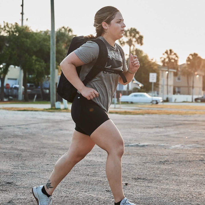A person jogging on a sunny day wears a gray T-shirt, GORUCK's Women’s Biker Shorts - ToughFlex featuring stay-put technology, and a backpack. A tattoo is visible on their leg, with trees and parked cars in the background.