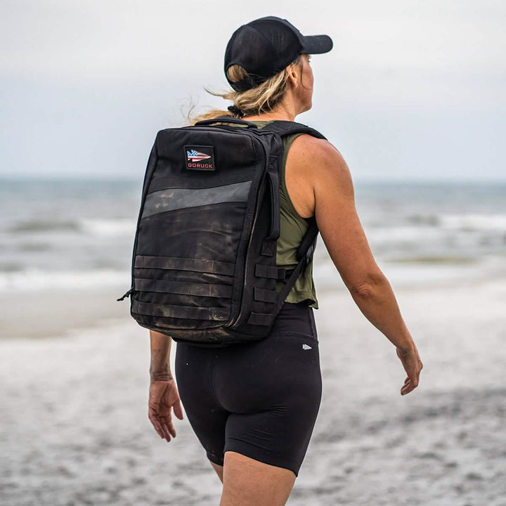A person sporting GORUCK’s Women's Biker Shorts in ToughFlex fabric strolls along a beach, with their back to the camera. They carry a black backpack and wear a cap, with the ocean and an overcast sky visible behind them, hinting at an adventure as resilient as their gear.