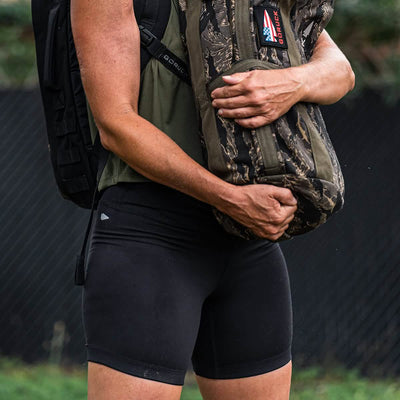 A person wearing GORUCK's ToughFlex Women’s Biker Shorts and a dark green top holds a camouflage-patterned bag adorned with an American flag patch. The backdrop features a black fence and lush greenery, accentuating the ensemble's rugged appeal.