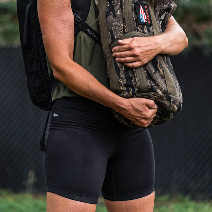 A person wearing GORUCK's ToughFlex Women’s Biker Shorts and a dark green top holds a camouflage-patterned bag adorned with an American flag patch. The backdrop features a black fence and lush greenery, accentuating the ensemble's rugged appeal.