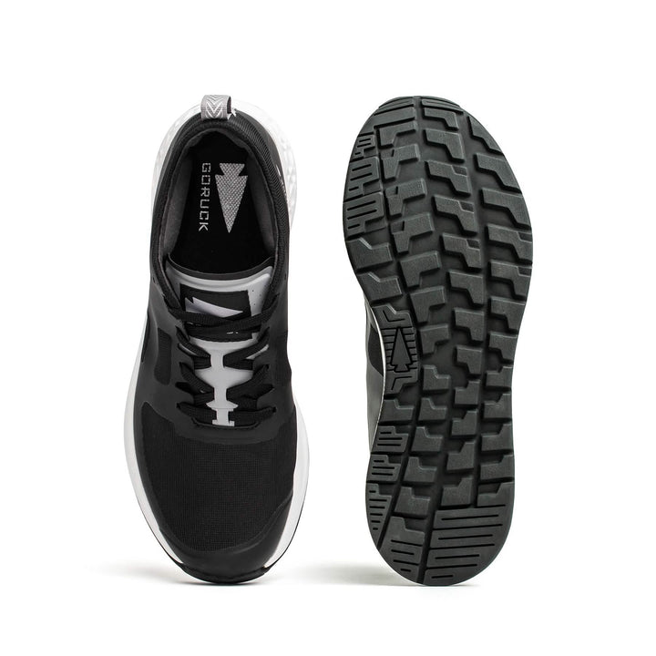 The Women's Rough Runner - Black + White sneakers from GORUCK are shown in both top and bottom views. The left shoe highlights the upper design with laces, while the right shoe displays the signature Gradient Density™ tread pattern on the sole, making them ideal for any Rough Runner™ adventure.