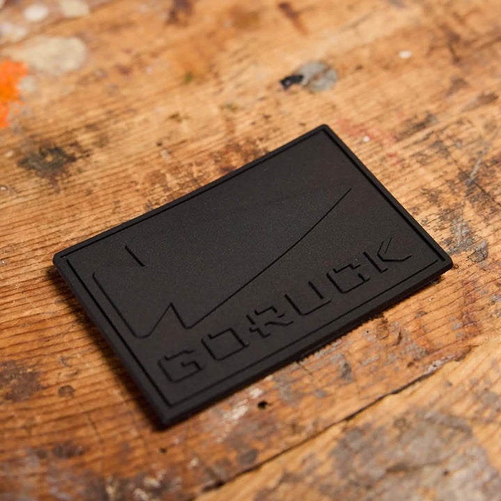 A black rectangular PVC patch from GORUCK displays the iconic spearhead design embossed alongside a knife, set against a textured wooden surface.