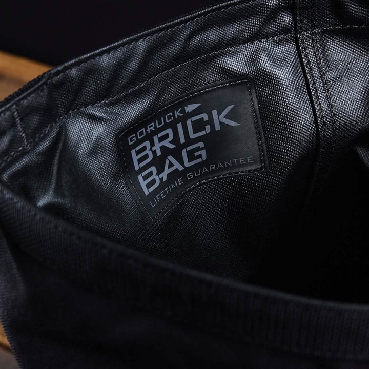 Close-up of a black fabric bag featuring a label that reads "GORUCK Brick Bag (5-50 LBS) - Lifetime Guarantee." The interior stitching and texture are inspired by military gear, providing versatile training utility.
