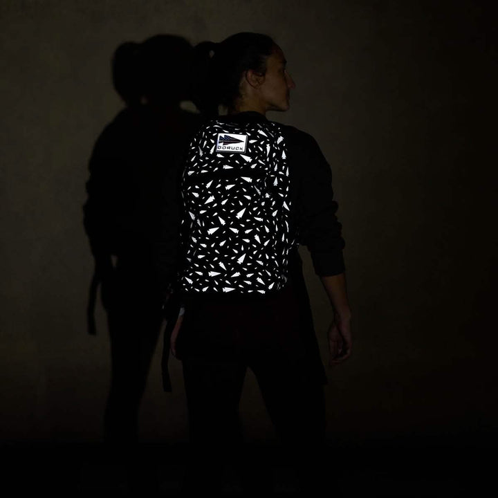 In front of a dark backdrop, an individual stands with the Bullet Ruck - Classic - Reflective Spearhead by GORUCK on their back, showcasing its durable Cordura construction and eye-catching reflective pattern that shines brightly. Casting a shadow on the wall as they face away, this backpack is designed for high visibility and is ideal for any adventure.