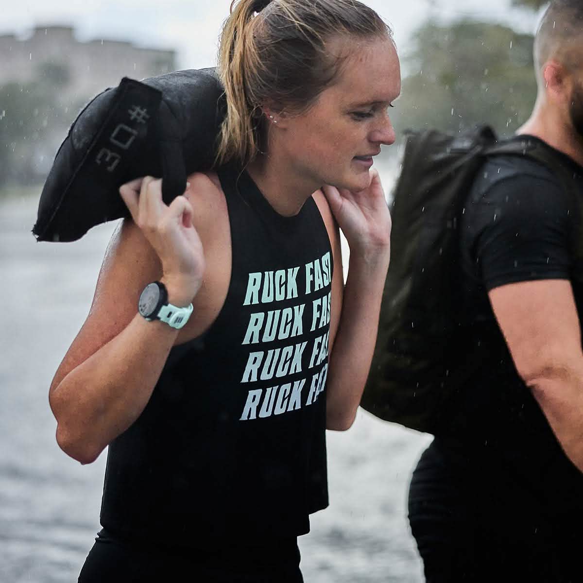 A woman strides through the rain, a burden on her shoulder, showcasing her motto "RUCK FAST" displayed on her Women’s Ruck Fast Tank - Tri-Blend by vendor-unknown. Raindrops cascade around her as she checks the watch on her wrist, determination evident in every step.