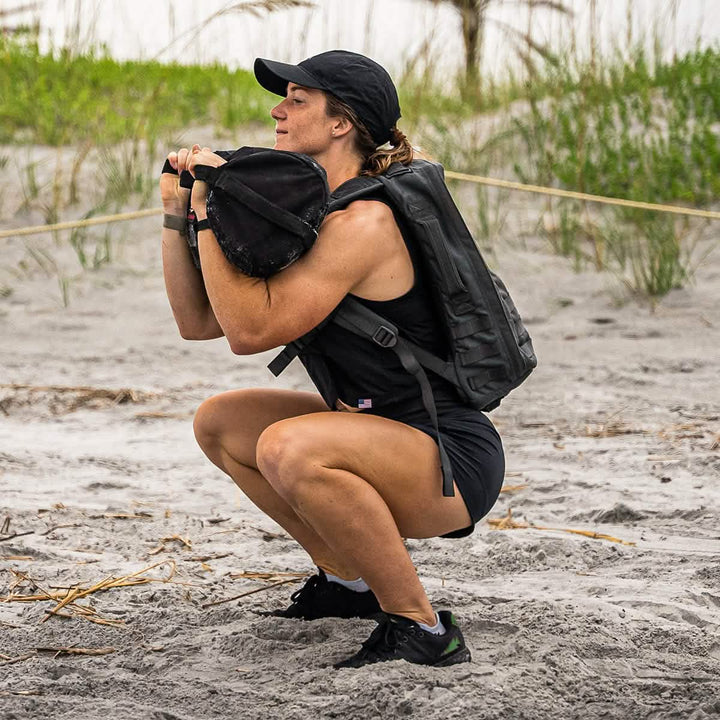 A person squats on a sandy beach, wearing ToughStretch athletic gear from GORUCK, complete with a black cap and a large backpack. They hold a weighted item to their chest while maintaining balance on the sand, where beachgrass sways gently against the blurred backdrop.