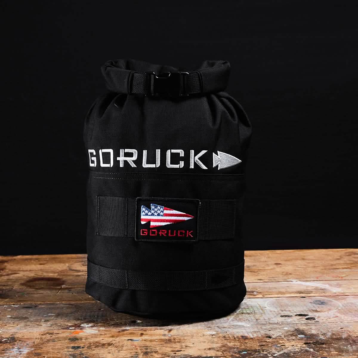 A black GORUCK Brick Bag (5-50 LBS), reminiscent of military gear, is placed on a wooden surface against a black background. It features a rolled top and displays a patch with an American flag design along with the brand name beneath it, emphasizing its versatile training capabilities.