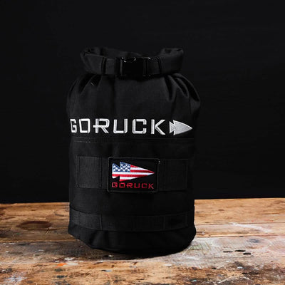A black GORUCK Brick Bag (5-50 LBS), reminiscent of military gear, is placed on a wooden surface against a black background. It features a rolled top and displays a patch with an American flag design along with the brand name beneath it, emphasizing its versatile training capabilities.