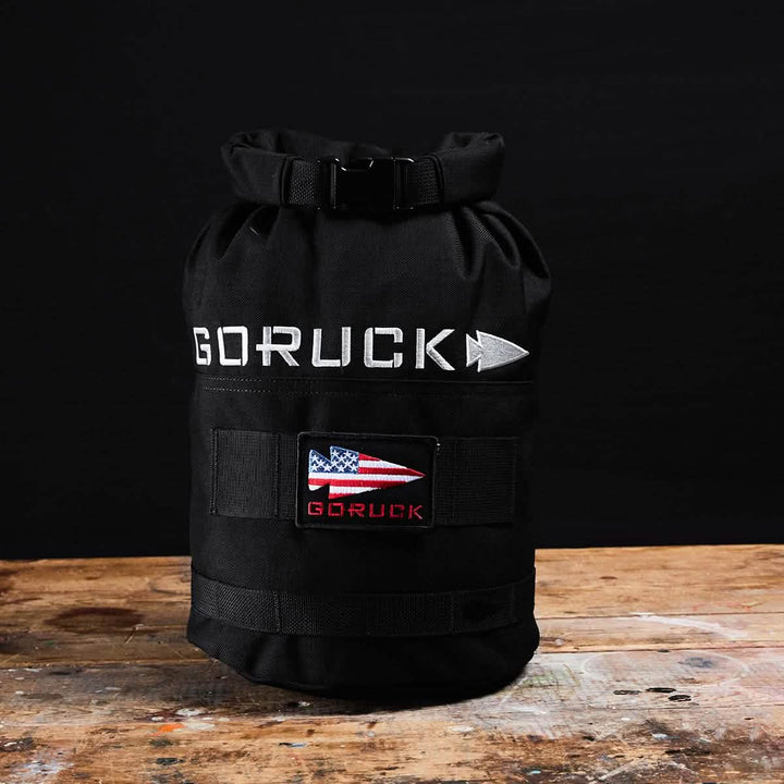 A black GORUCK Brick Bag (5-50 LBS), reminiscent of military gear, is placed on a wooden surface against a black background. It features a rolled top and displays a patch with an American flag design along with the brand name beneath it, emphasizing its versatile training capabilities.