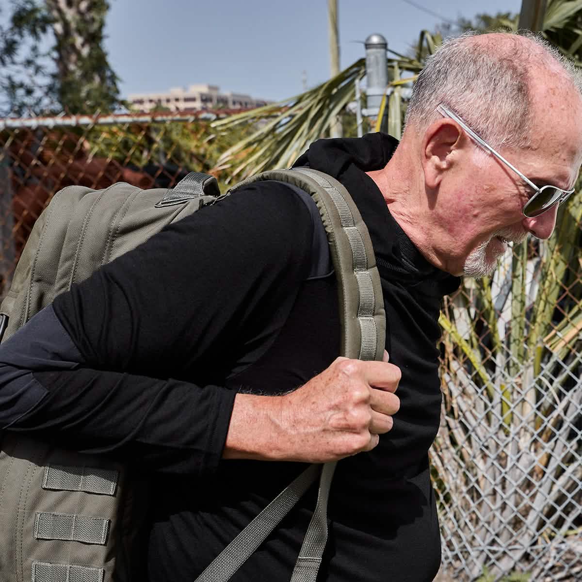 An older man with short white hair and a beard, accessorized with sunglasses and donning the GORUCK Men’s Commando Pullover made of Merino wool, carries a large backpack. He walks leisurely outdoors past a chain-link fence with trees in the background.