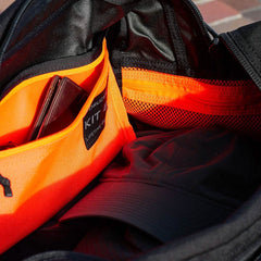 A Kit Bag from GORUCK, reminiscent of a Special Forces Flight Bag, reveals an orange inner compartment containing a wallet and a black cap. Sunlight highlights the contrast between the black interior and the bright orange pocket.