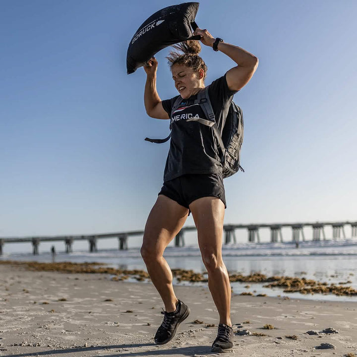 On a sandy beach, someone demonstrates their squat-proof **Women’s Training Shorts - ToughStretch** from GORUCK by lifting a weighted bag overhead. They are wearing a backpack and a dark shirt featuring "America," with a bridge and the calm sea providing the backdrop under a clear blue sky.