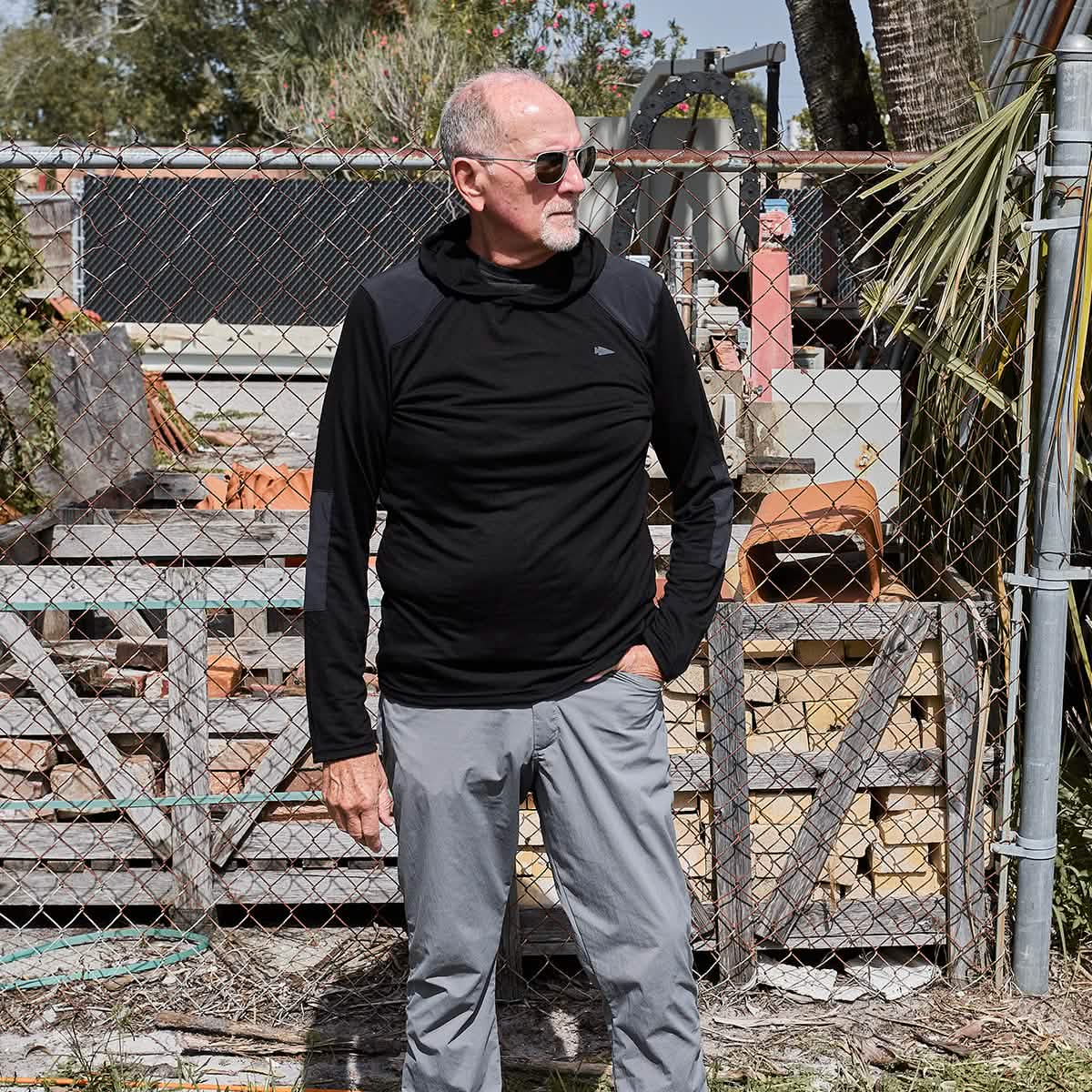 A man with a bald head and sunglasses is standing outside in front of a chain-link fence, wearing a stylish black Men’s Commando Pullover made from merino wool by GORUCK, paired with gray pants. The background features wooden pallets and various outdoor items.