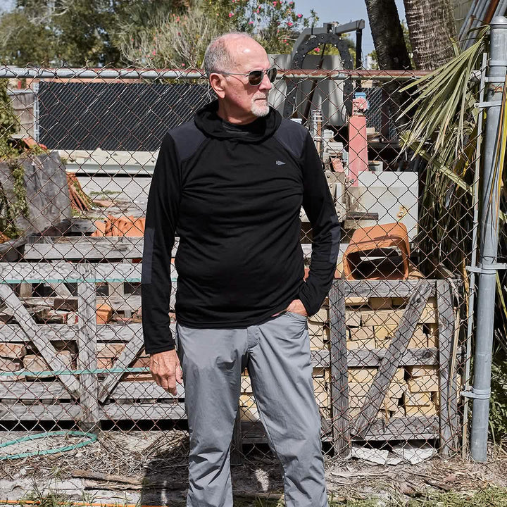 A man with a bald head and sunglasses is standing outside in front of a chain-link fence, wearing a stylish black Men’s Commando Pullover made from merino wool by GORUCK, paired with gray pants. The background features wooden pallets and various outdoor items.