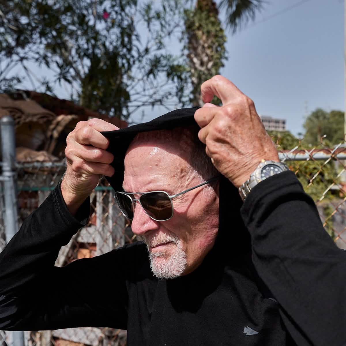 An elderly man wearing sunglasses adjusts his GORUCK Men’s Commando Pullover made of merino wool outdoors. A chain-link fence and trees can be seen in the background on a sunny day.