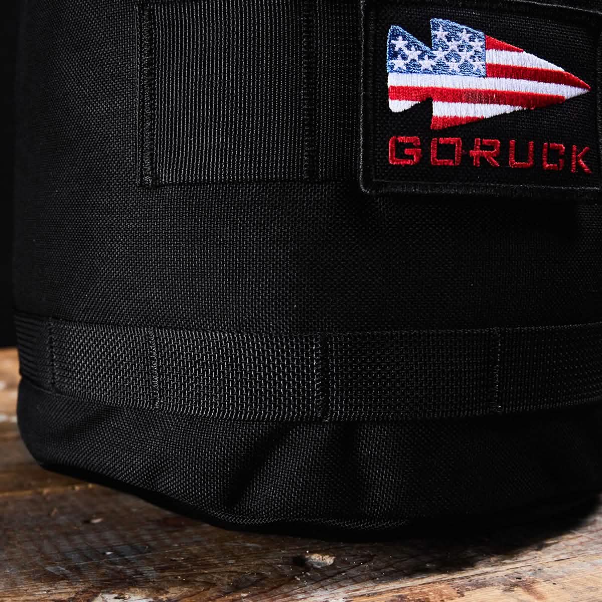 A close-up shot of the Brick Bag (5-50 LBS), a black fabric bag from GORUCK, designed as versatile training gear. It features a patch showcasing a spearhead design with the American flag, with "GORUCK" embroidered in red beneath it. Resting on a wooden surface, this military gear bag embodies durability and style.